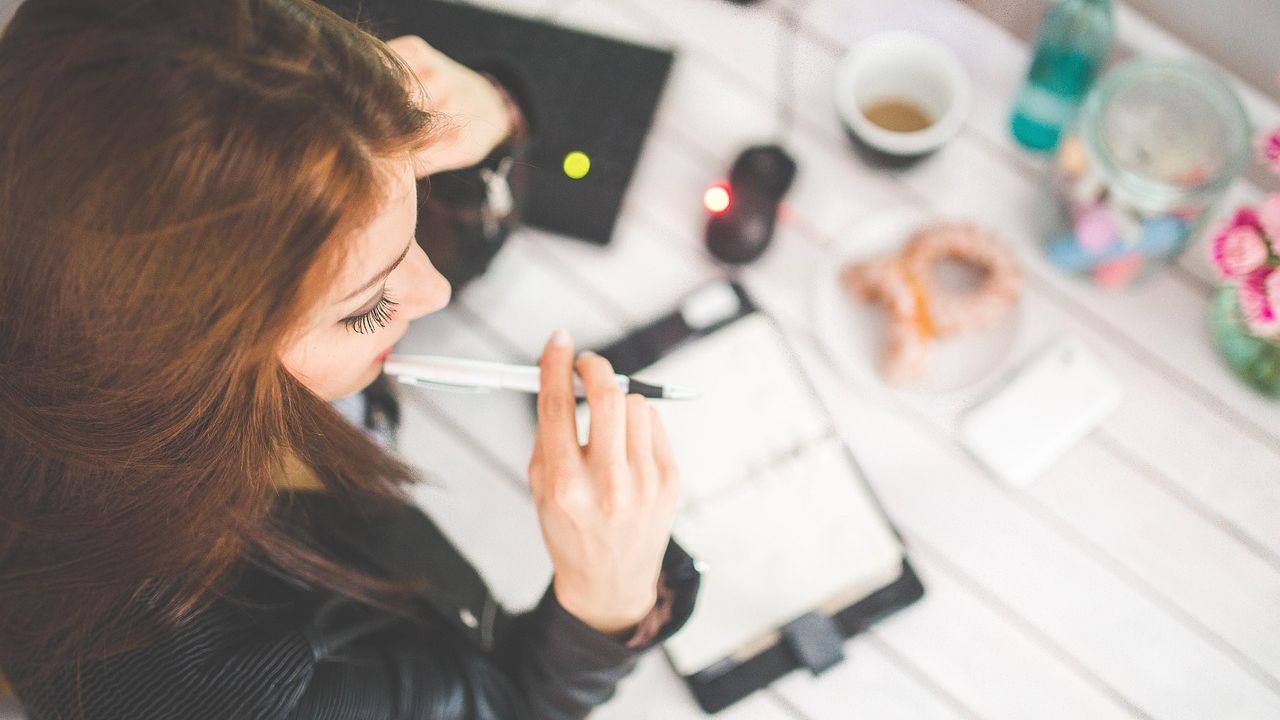 Ảnh chụp từ trên xuống: một người cầm bút bên sổ tay trên bàn trắng, cạnh tách cà phê và điện thoại