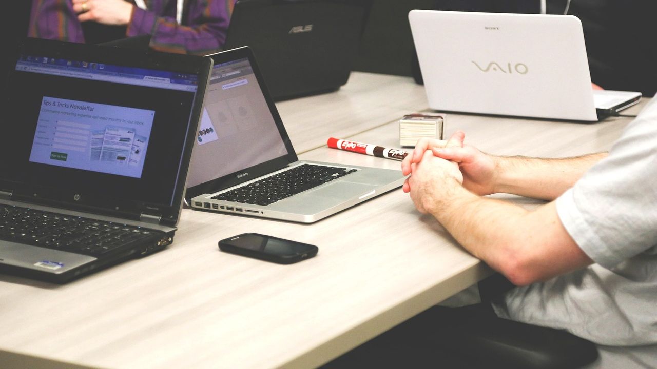 Ảnh chụp bàn họp với nhiều laptop đang mở, một người ngồi khoanh tay theo dõi màn hình