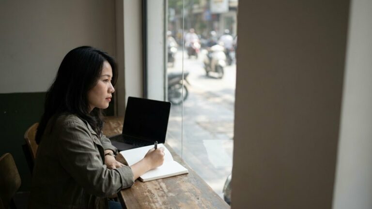 Người phụ nữ ngồi quầy cửa sổ quán cà phê, máy tính xách tay mở và cuốn sổ trước mặt, đường phố mờ ngoài kính