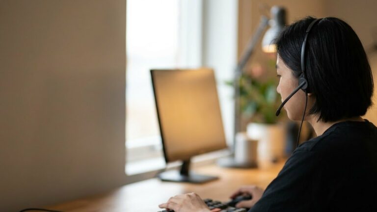 Nữ nhân viên đeo tai nghe ngồi trước màn hình máy tính, tay đặt trên bàn phím, đèn bàn và cây nhỏ phía sau