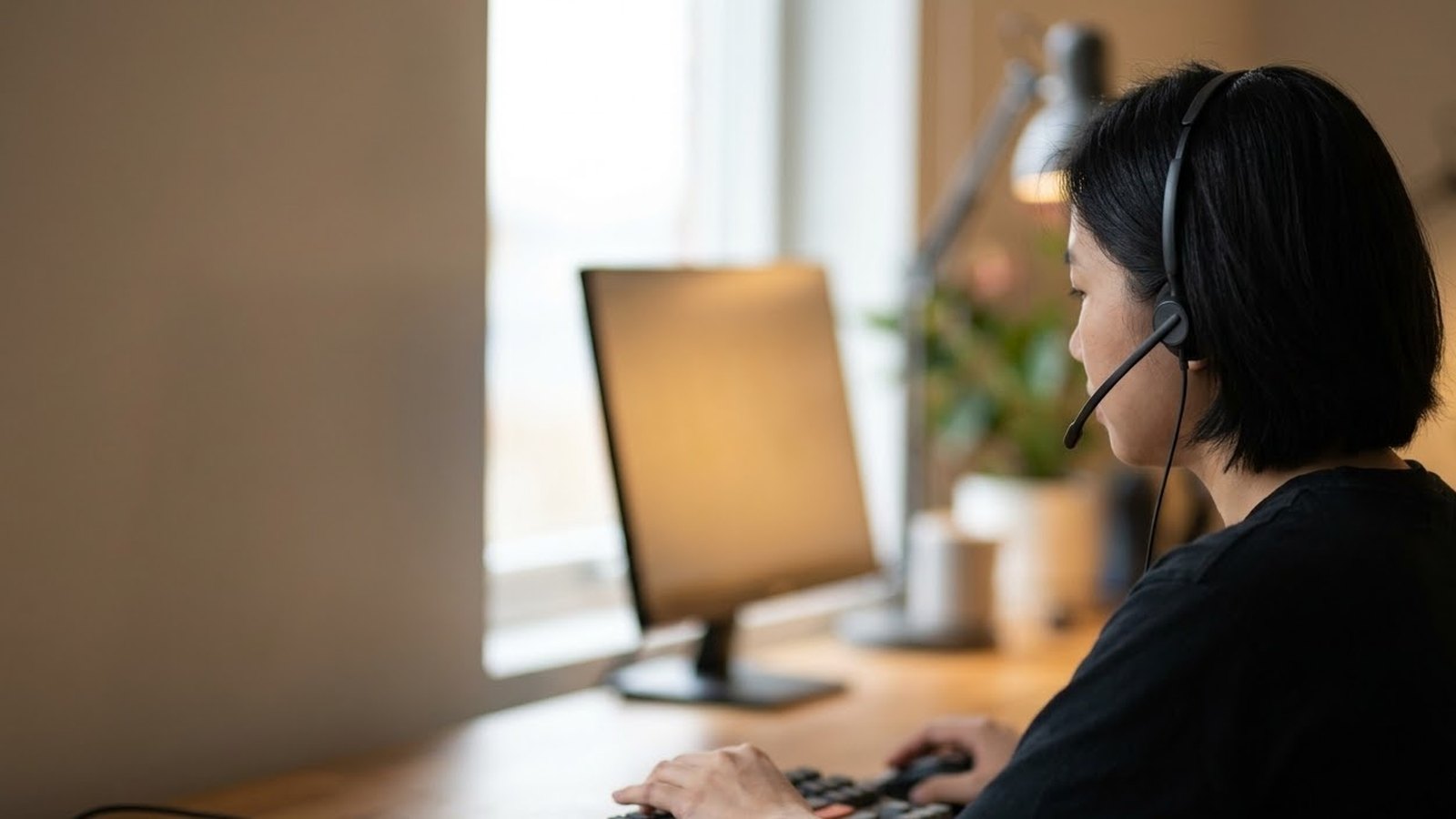 Nữ nhân viên đeo tai nghe ngồi trước màn hình máy tính, tay đặt trên bàn phím, đèn bàn và cây nhỏ phía sau