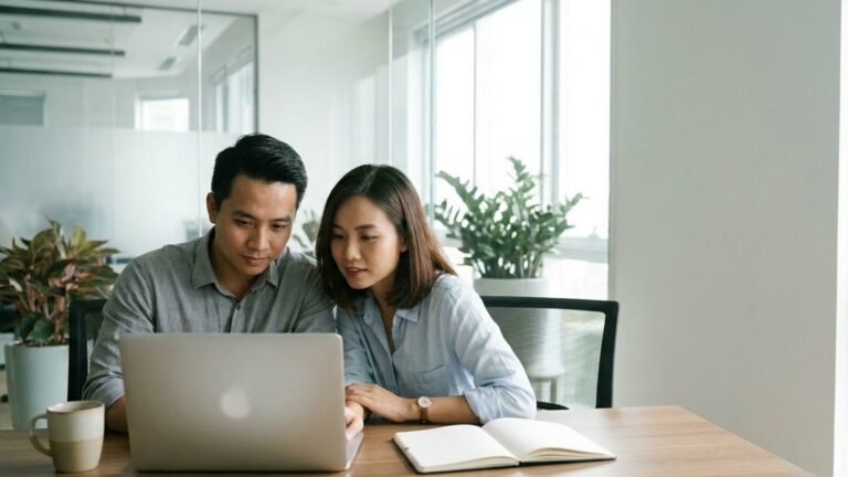 Hai đồng nghiệp ngồi cạnh nhau tại bàn làm việc cùng nhìn màn hình laptop, sổ tay mở và cốc cà phê bên cạnh, văn phòng sáng nhiều cây xanh