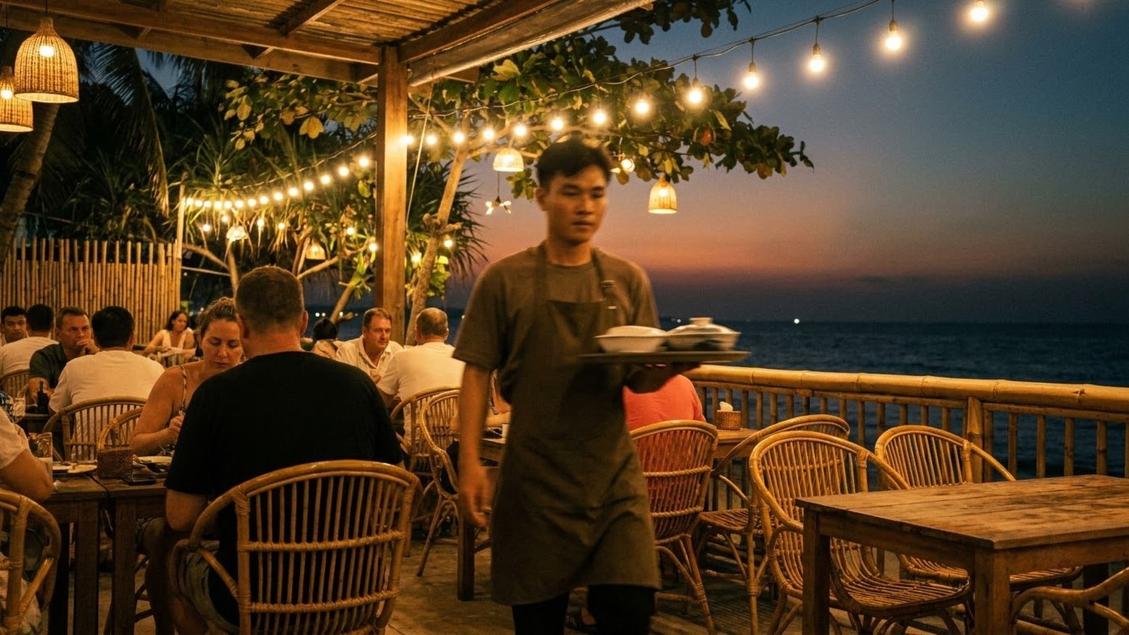 A seaside restaurant terrace at dusk, a server carrying a tray between occupied tables