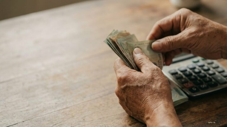 Cận cảnh đôi bàn tay lớn tuổi đếm xấp tiền giấy bên chiếc máy tính bỏ túi đặt trên mặt bàn gỗ mộc